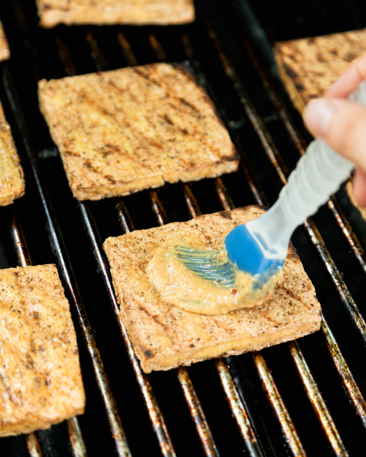 Μια εικόνα γωνίας 3/4 δείχνει φέτες tofu στη σχάρα σε μια σχάρα. Μια φέτα βουρτσίζεται με κρεμώδη σάλτσα.